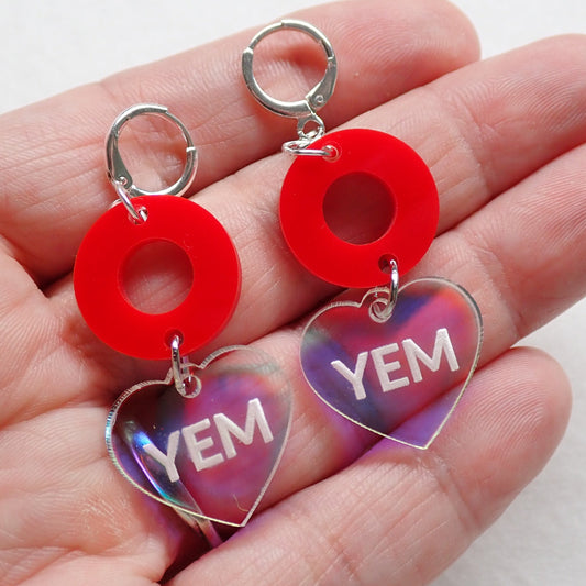 Red heart-shaped earrings with 'YEM' text held in a hand against a white background